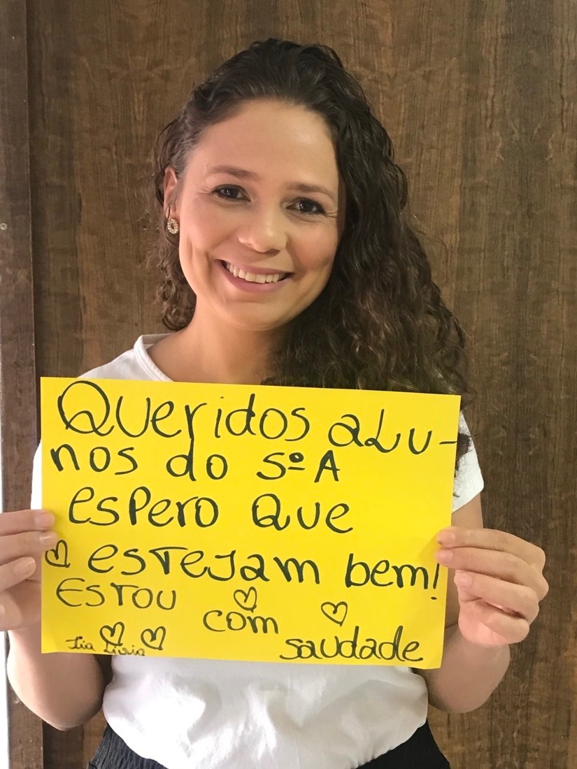 Lívia segurando um cartaz amarelo: Queridos alunos do 5º A, espero que estejam bem! Estou com saudade.